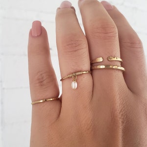 May include: Three gold rings on a hand. The ring on the middle finger has a small pearl dangling from it.