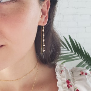 May include: A gold threader earring featuring a delicate chain adorned with five small, round, white pearls. The earring is worn through the earlobe, with the chain hanging vertically, showcasing a minimalist design with a polished gold finish.