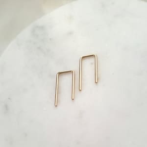 May include: Two gold-colored, minimalist U-shaped earrings. The earrings are placed on a white marble surface. The earrings have a simple, geometric design, with a rectangular shape.