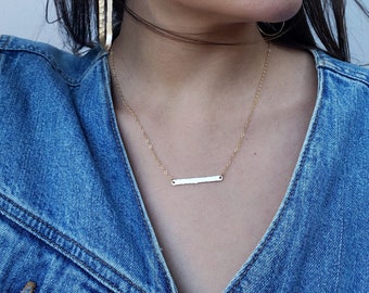 May include: A gold bar necklace and matching gold earrings are displayed. The earrings have a textured, vertical design. The necklace features a simple, horizontal bar pendant. The model is wearing a denim jacket.