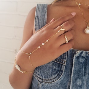 May include: A gold chain bracelet with pearl accents and a larger pearl charm. The bracelet is worn on the wrist and extends up the arm, ending in a ring on the index finger.