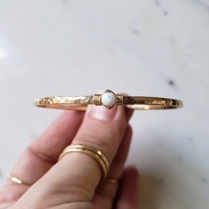 May include: A gold hammered bangle bracelet with a white opal gemstone set in the center.
