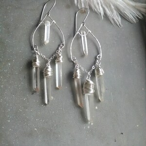 May include: Silver wire wrapped earrings with clear quartz crystals dangling from a silver hoop. The crystals are wrapped in silver wire and hang in a cascading design.