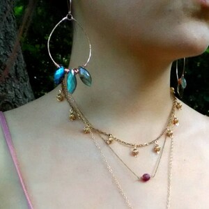 May include: Gold-toned jewelry set featuring large hoop earrings with blue-green gemstone accents and a layered necklace. The necklace includes small gold charms and a single, round, red gemstone pendant. The jewelry is worn against a light pink top.