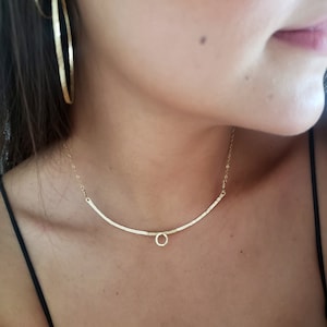 May include: A gold necklace with a delicate, curved bar design and a small, circular pendant. The necklace is worn by a person with dark hair.