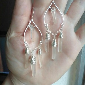 May include: Silver wire wrapped earrings with clear quartz crystal drops. The earrings feature a teardrop shape with a single crystal at the top and three crystals dangling from a chain below.