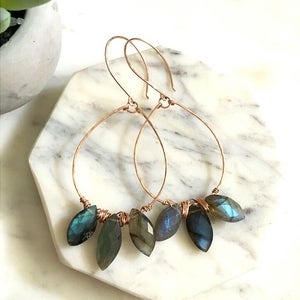 May include: Copper wire hoop earrings with teardrop-shaped labradorite stones. The stones are a mix of blues, greens, and grays. The earrings are displayed on a white and gray marble surface.