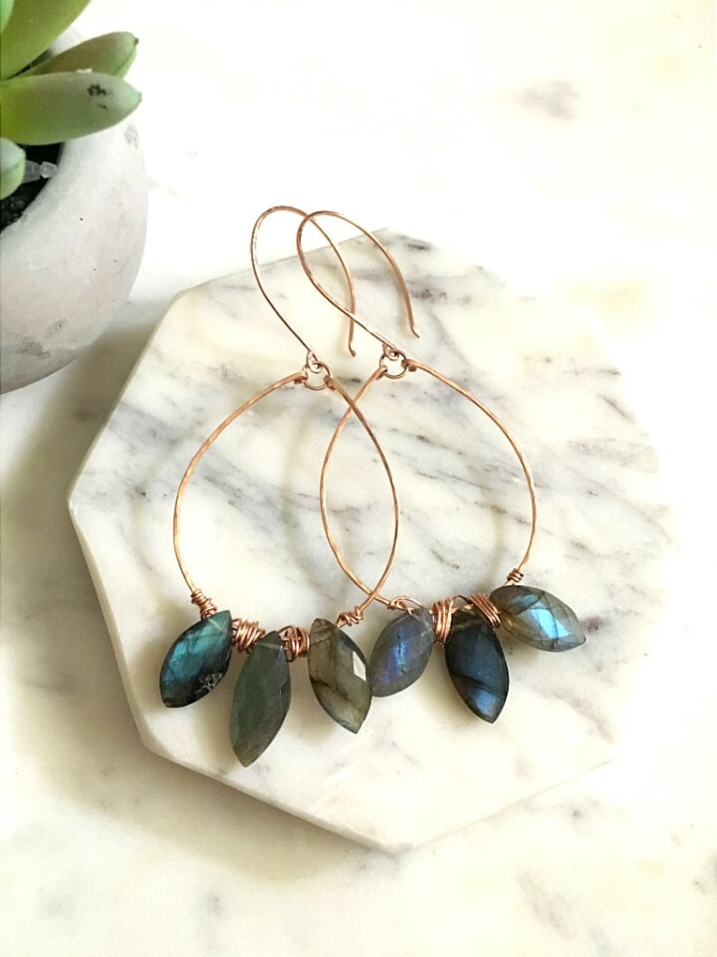 May include: Copper wire hoop earrings with teardrop-shaped labradorite stones. The stones are a mix of blues, greens, and grays. The earrings are displayed on a white and gray marble surface.