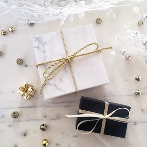 May include: Two gift boxes wrapped in white marble paper and black paper with gold ribbon. The boxes are on a white marble surface with gold and silver decorative accents.