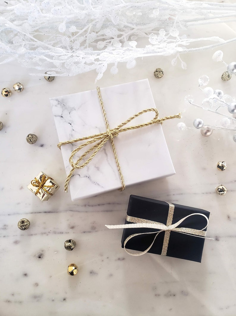 May include: Two gift boxes wrapped in white marble paper and black paper with gold ribbon. The boxes are on a white marble surface with gold and silver decorative accents.