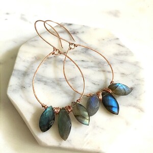 May include: Copper wire hoop earrings with teardrop-shaped, iridescent blue-green gemstones. The earrings are displayed on a white and gray marble surface. The gemstones are wire-wrapped to the bottom of the hoops.