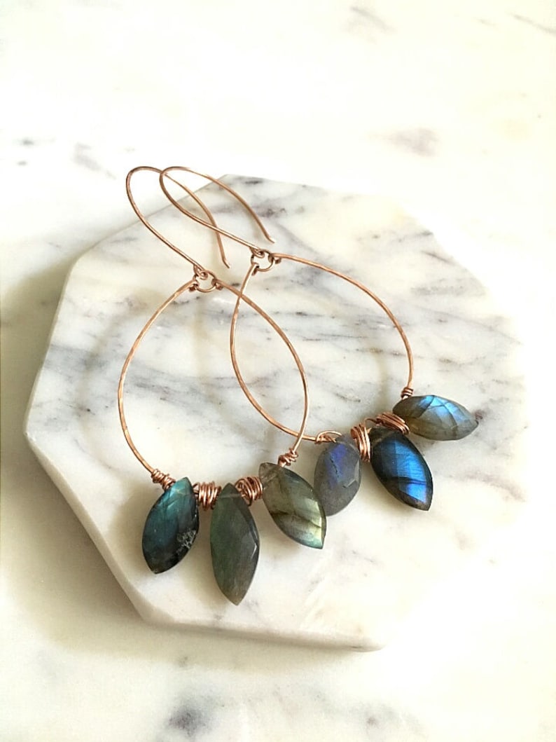 May include: Copper wire hoop earrings with teardrop-shaped, iridescent blue-green gemstones. The earrings are displayed on a white and gray marble surface. The gemstones are wire-wrapped to the bottom of the hoops.