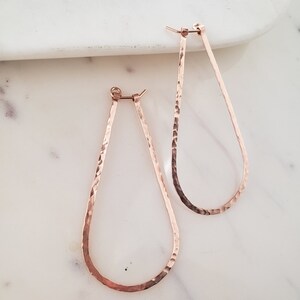 Puede incluir: Un par de pendientes de aro de l&aacute;grima martillados en oro rosa. Los pendientes est&aacute;n hechos de un metal delgado y tienen una textura martillada.