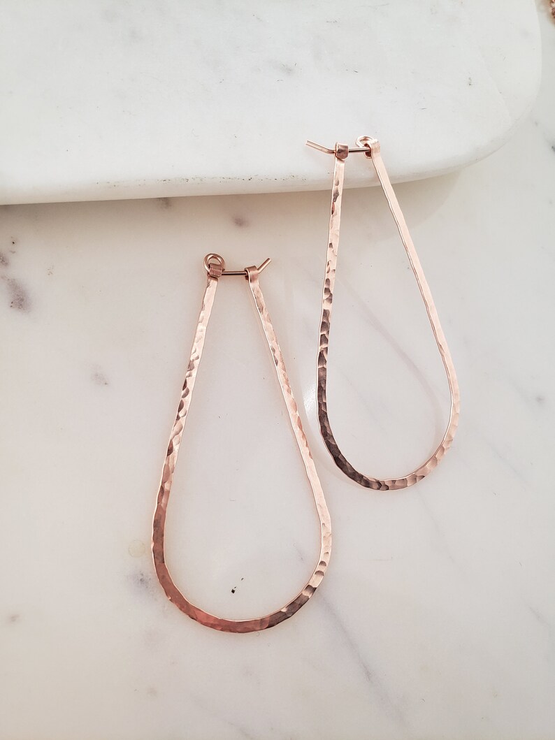 Puede incluir: Un par de pendientes de aro de l&aacute;grima martillados en oro rosa. Los pendientes est&aacute;n hechos de un metal delgado y tienen una textura martillada.