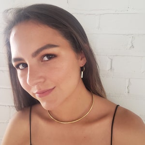 Puede incluir: Una mujer lleva un collar choker dorado y pendientes de aro dorados con un peque&ntilde;o colgante de barra. Lleva una camiseta de tirantes negra.