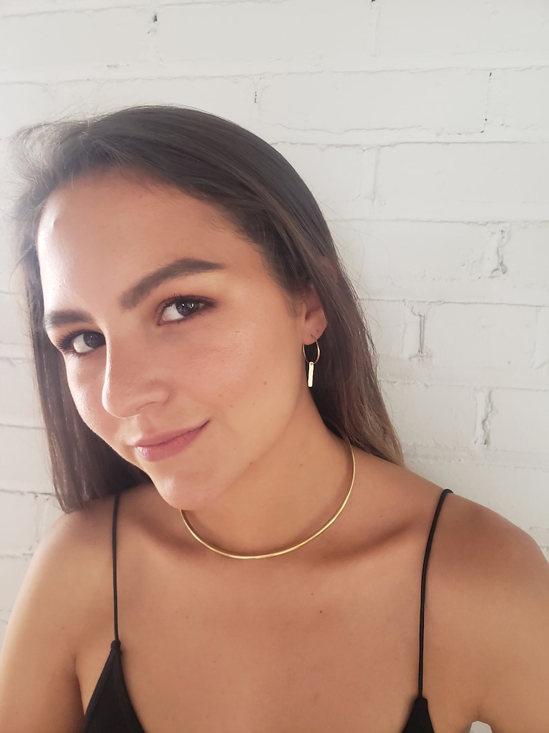 Puede incluir: Una mujer lleva un collar choker dorado y pendientes de aro dorados con un peque&ntilde;o colgante de barra. Lleva una camiseta de tirantes negra.