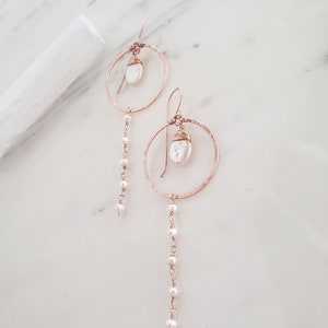 May include: A pair of rose gold hoop earrings with a pearl dangle. Each earring features a hammered hoop with a pearl charm and a delicate chain of pearls hanging below.