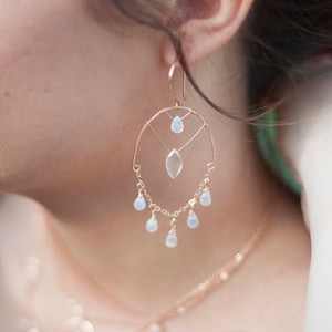May include: A rose gold hoop earring featuring a wire-wrapped frame with a central marquise-cut clear stone, a small teardrop stone above it, and five dangling teardrop-shaped moonstone accents suspended from the bottom of the hoop.