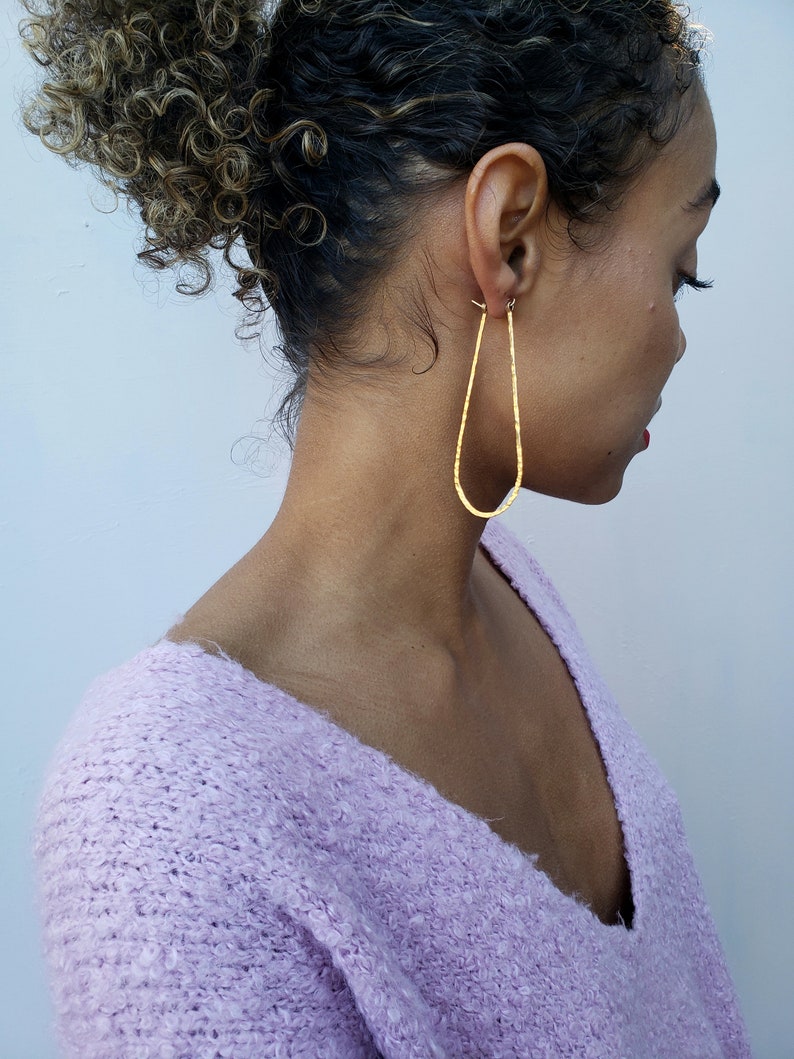 Puede incluir: Un par de pendientes de oro en forma de l&aacute;grima con textura martillada. Los pendientes son usados por una persona con cabello rizado.