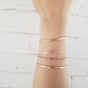 May include: A set of four gold bracelets stacked on a person's wrist. The bracelets are all hammered and have a slightly wavy shape.
