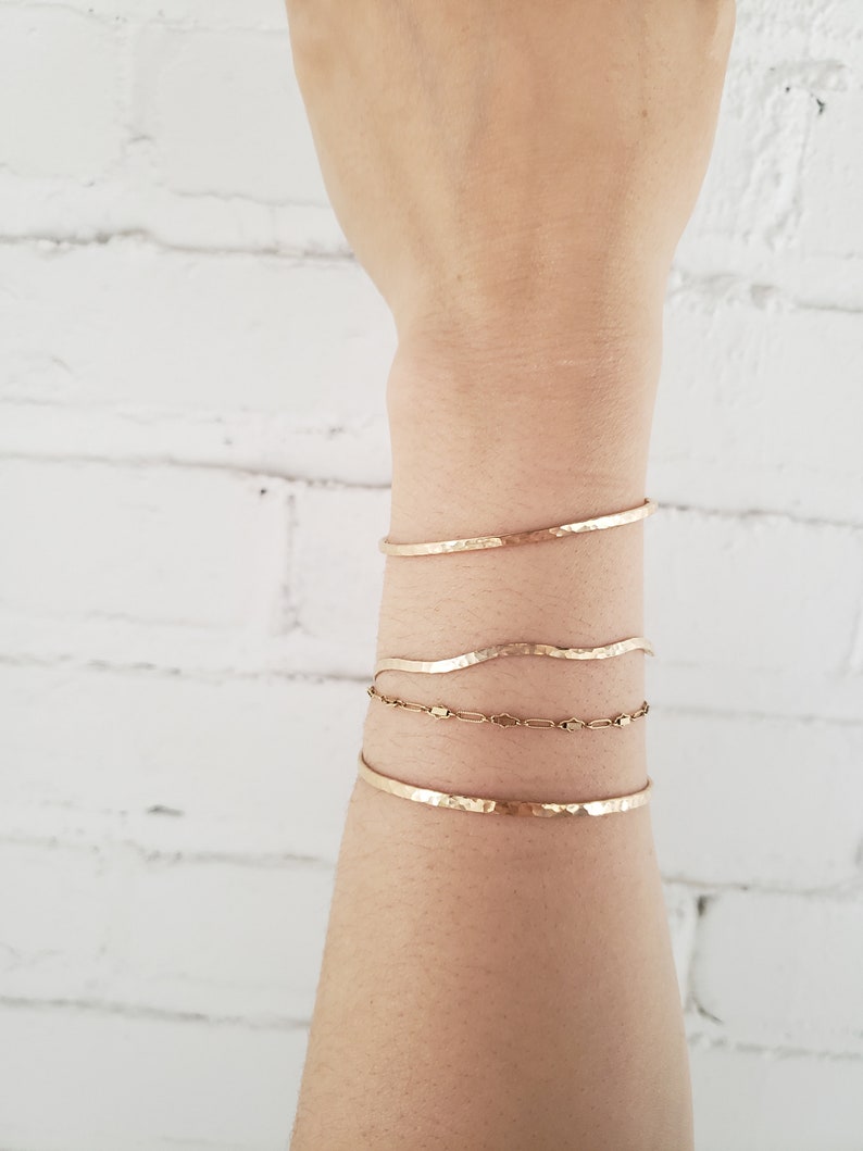May include: A set of four gold bracelets stacked on a person's wrist. The bracelets are all hammered and have a slightly wavy shape.