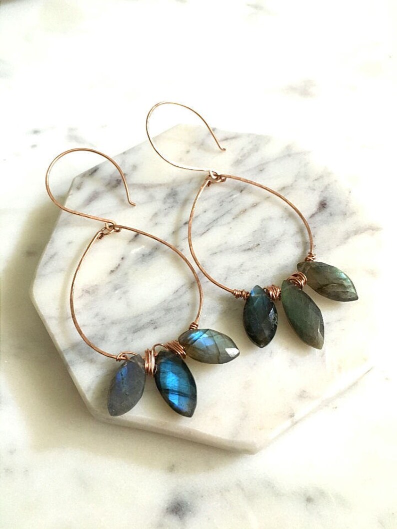 May include: A pair of copper-toned hoop earrings. Each earring features a teardrop shape with dangling, iridescent blue-green gemstone beads. The earrings are displayed on a white and gray marble surface.