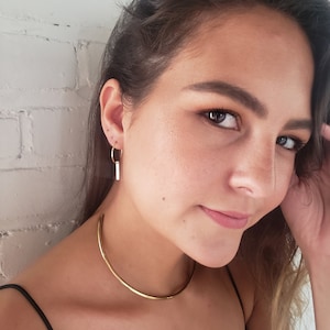 Puede incluir: Una mujer con un collar de gargantilla de oro y pendientes de aro de oro con un colgante de barra vertical.