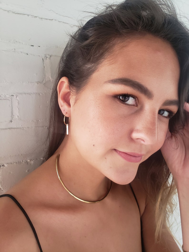 Puede incluir: Una mujer con un collar de gargantilla de oro y pendientes de aro de oro con un colgante de barra vertical.