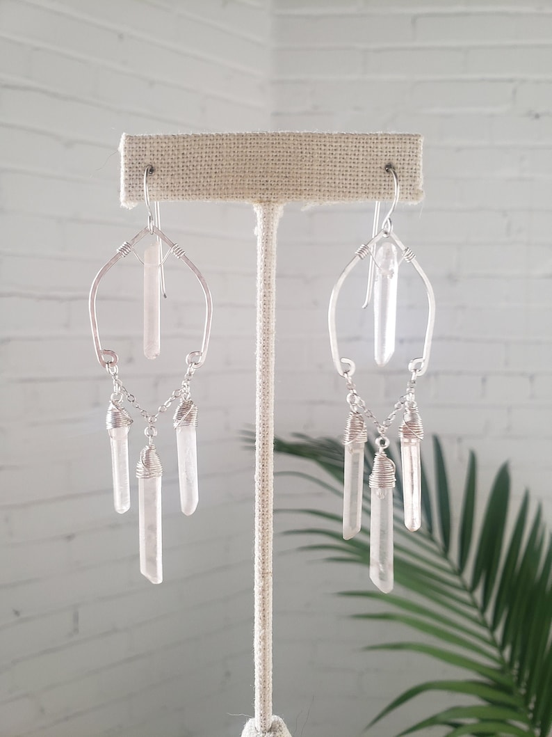 May include: Silver wire wrapped earrings with clear quartz crystals. The earrings feature a long crystal dangling from the top and three smaller crystals dangling from the bottom.