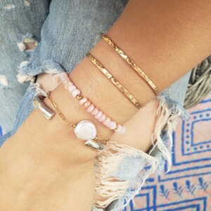 May include: A set of three gold hammered bracelets stacked on a wrist, along with a delicate chain bracelet with a pearl charm and a pink beaded bracelet.