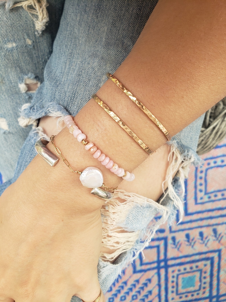 May include: A set of three gold hammered bracelets stacked on a wrist, along with a delicate chain bracelet with a pearl charm and a pink beaded bracelet.