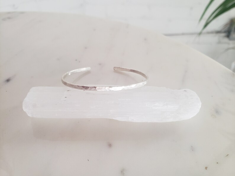 May include: A silver hammered bangle bracelet. The bracelet is thin and delicate, with a smooth, polished finish.