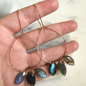 May include: Copper hoop earrings with faceted, teardrop-shaped labradorite stones. The stones display iridescent blue and green hues. The hoops are hammered for texture and have a simple hook closure.