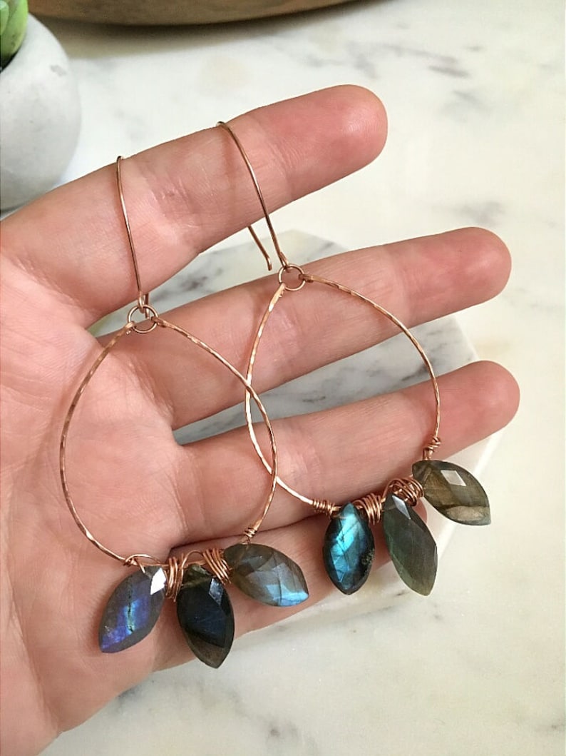 May include: Copper hoop earrings with faceted, teardrop-shaped labradorite stones. The stones display iridescent blue and green hues. The hoops are hammered for texture and have a simple hook closure.