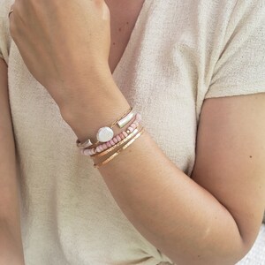 May include: A woman's wrist wearing a stack of bracelets, including a silver bangle with a pearl charm, a pink beaded bracelet, and two gold bracelets.