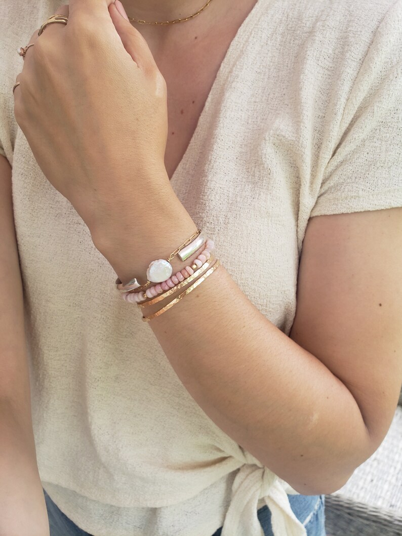 May include: A woman's wrist wearing a stack of bracelets, including a silver bangle with a pearl charm, a pink beaded bracelet, and two gold bracelets.