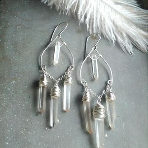 May include: Silver dangle earrings with clear quartz crystals wrapped in wire. The earrings feature a teardrop shape with a single crystal at the top and three crystals dangling below.