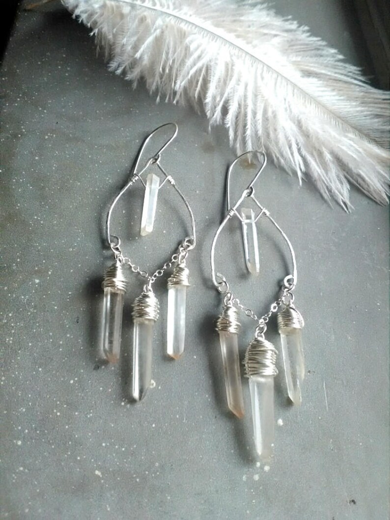 May include: Silver dangle earrings with clear quartz crystals wrapped in wire. The earrings feature a teardrop shape with a single crystal at the top and three crystals dangling below.