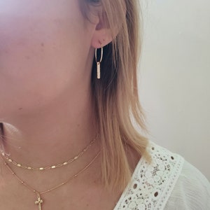Puede incluir: Una mujer lleva una blusa blanca con un delicado ribete de encaje, pendientes de aro dorados con un colgante de barra y un collar de oro en capas con un peque&ntilde;o colgante de cruz.