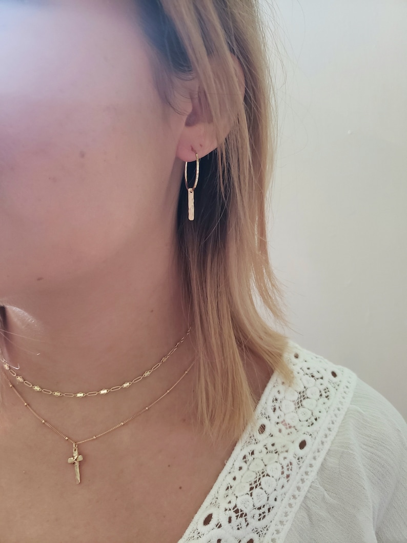 Puede incluir: Una mujer lleva una blusa blanca con un delicado ribete de encaje, pendientes de aro dorados con un colgante de barra y un collar de oro en capas con un peque&ntilde;o colgante de cruz.