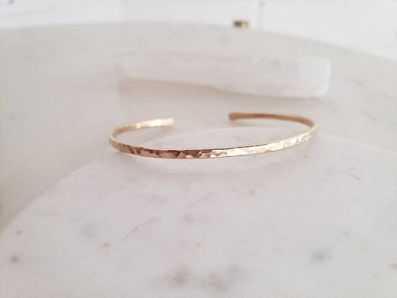 May include: A delicate gold hammered bangle bracelet. The bracelet is thin and has a smooth, polished finish.