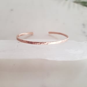 May include: A hammered rose gold cuff bracelet. The bracelet is thin and delicate, with a smooth, polished finish.