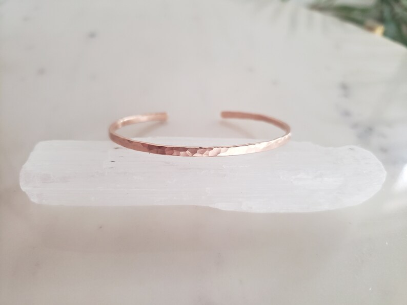 May include: A hammered rose gold cuff bracelet. The bracelet is thin and delicate, with a smooth, polished finish.