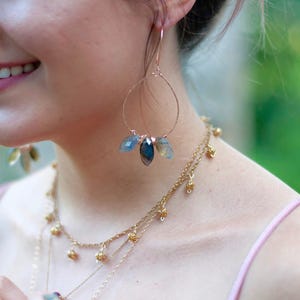 May include: Rose gold hoop earrings with three blue and gray gemstone pendants. A layered gold necklace with small decorative beads is also visible. The jewelry is worn with a pink spaghetti strap top.