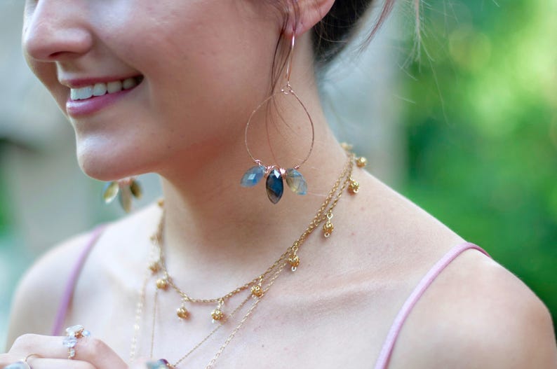May include: Rose gold hoop earrings with three blue and gray gemstone pendants. A layered gold necklace with small decorative beads is also visible. The jewelry is worn with a pink spaghetti strap top.