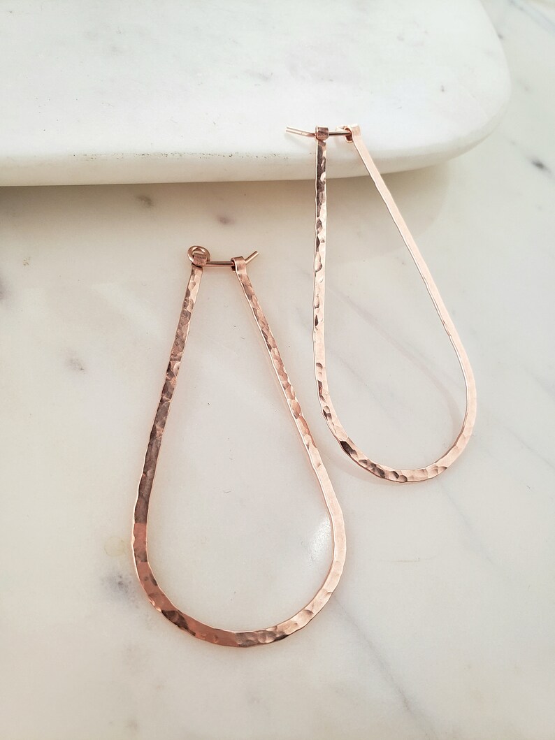 Puede incluir: Un par de pendientes de aro ovalados martillados en oro rosa. Los pendientes son grandes y tienen una textura martillada.