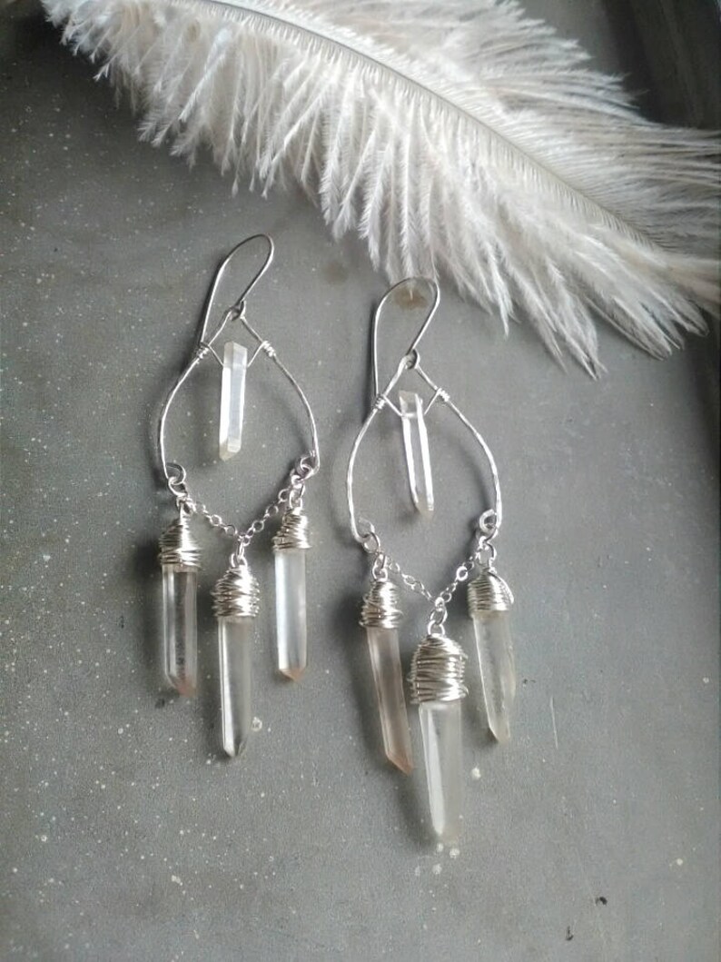 May include: Silver wire wrapped earrings with clear quartz crystals dangling from a silver hoop. The crystals are wrapped in silver wire and hang in a cascading design.