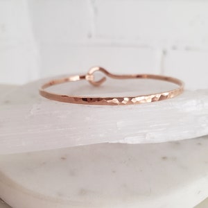 May include: A hammered rose gold bangle bracelet with a simple, open-circle design. The bracelet is made of a thin, delicate metal and has a slightly textured finish.