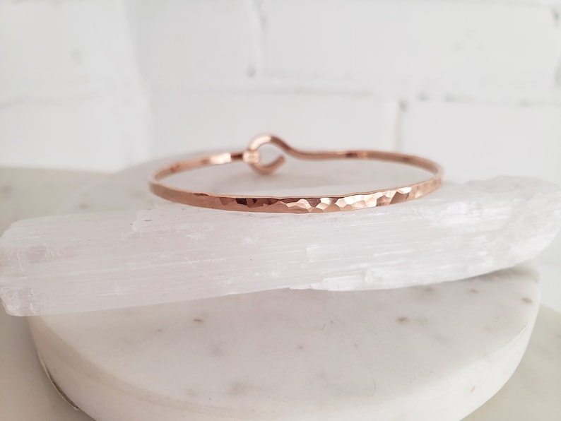 May include: A hammered rose gold bangle bracelet with a simple, open-circle design. The bracelet is made of a thin, delicate metal and has a slightly textured finish.