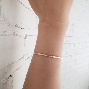 May include: A thin, hammered, rose gold bracelet on a person's wrist.